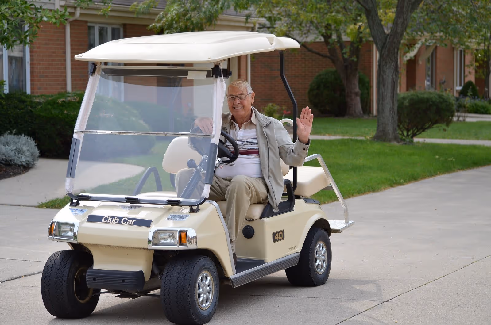 An elderly man wearing glasses and a light jacket is sitting in a beige golf cart on a paved path outside a brick building with green grass and trees in the background. He is smiling and waving with his right hand.