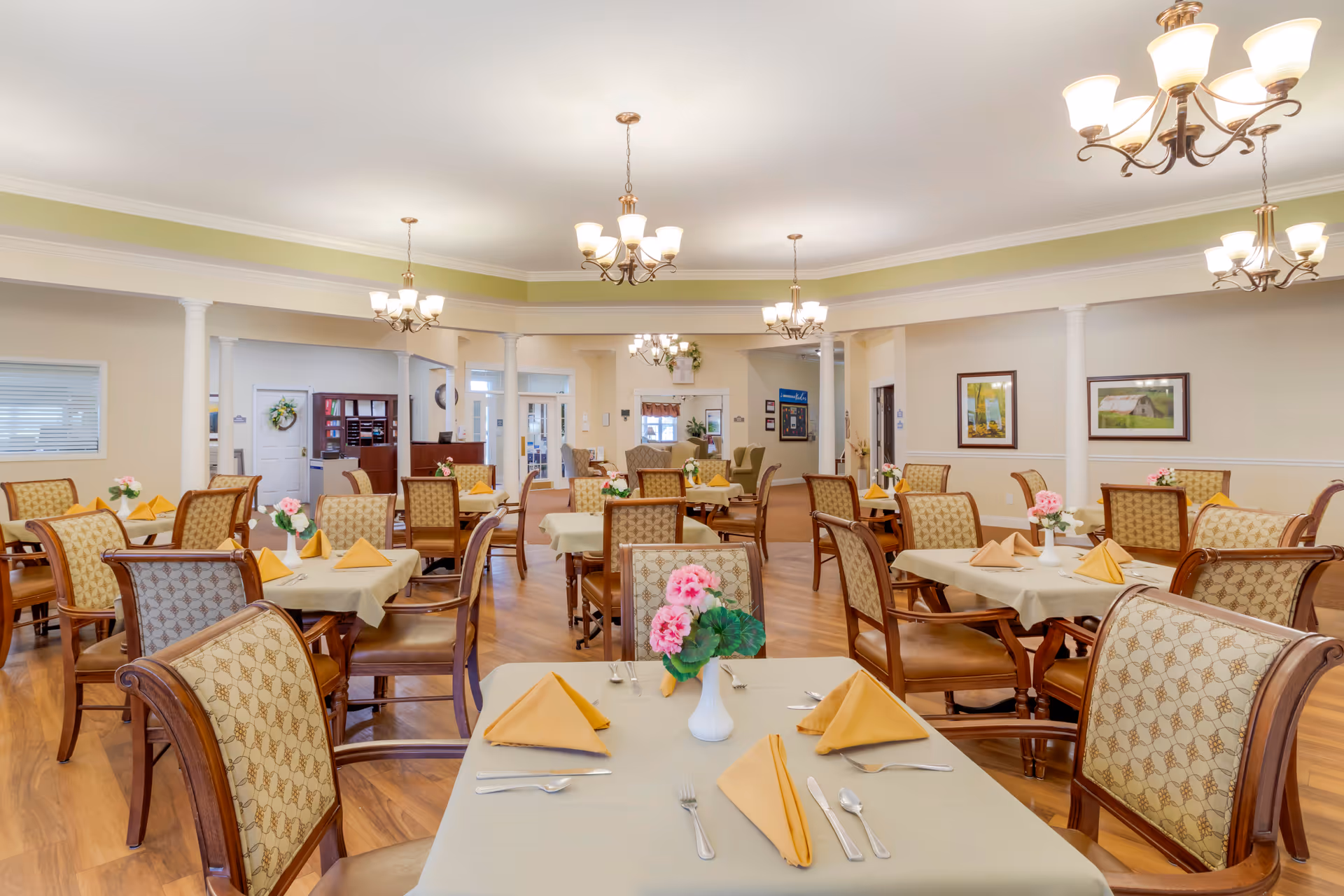 A spacious dining room in a senior living facility with multiple tables covered in beige tablecloths, each set with yellow folded napkins, silverware, and a white vase holding pink flowers. The room features wooden chairs with patterned upholstery, hardwood floors, cream-colored walls with white trim, decorative columns, framed artwork, and multiple chandeliers providing warm lighting.