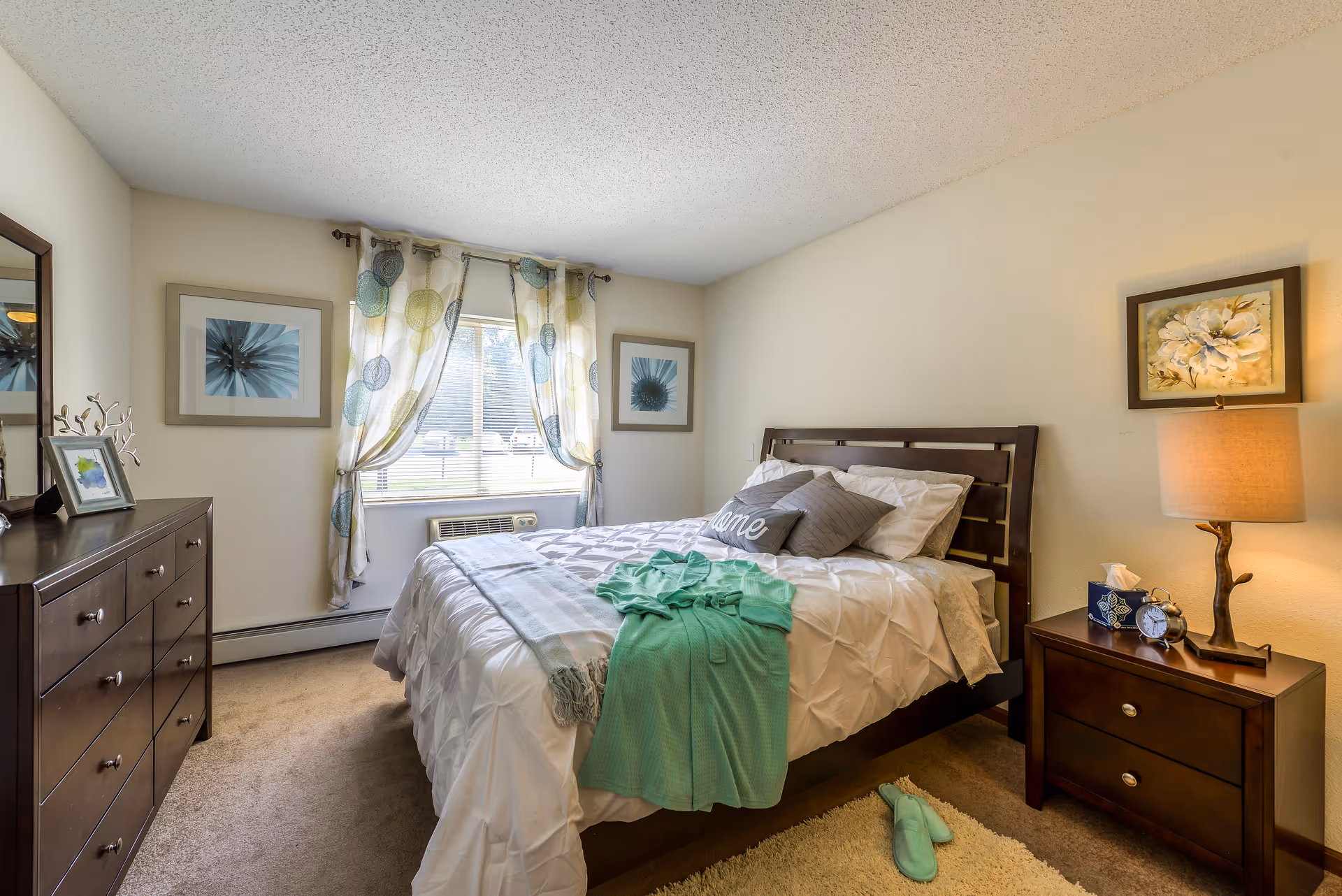 Neatly made bedroom with a wooden bed, matching nightstand and dresser, window with patterned curtains, and teal slippers on the floor.