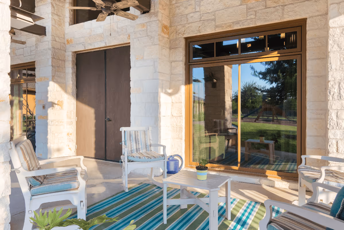 Outdoor patio area with white cushioned chairs and a small table on a striped blue and green rug. The patio is surrounded by stone walls and has a ceiling fan above. A large glass sliding door reflects the green lawn and trees outside.
