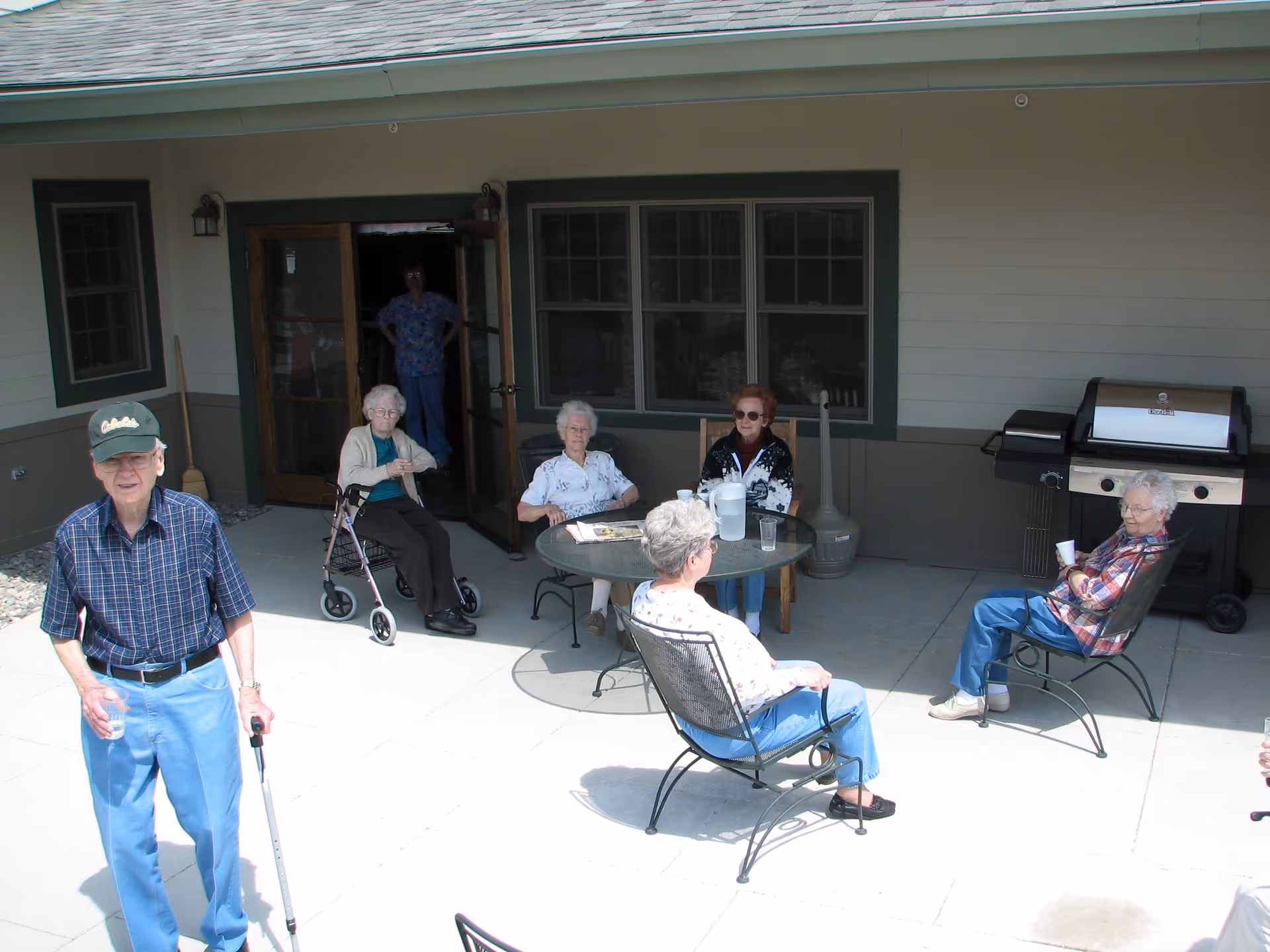 Several elderly residents sitting and socializing on a covered patio outside a senior living building, with a man standing in the foreground using a cane.