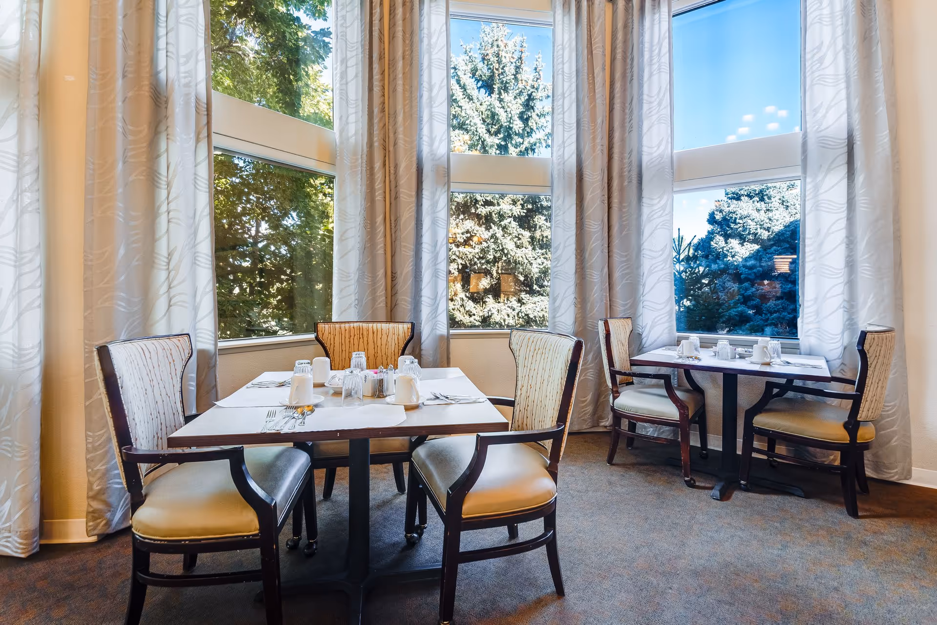 A bright dining area with two tables, each surrounded by four cushioned chairs. The tables are set with white cups, glasses, and silverware. Large windows with light-colored curtains allow natural light to fill the room and provide a view of green trees outside.