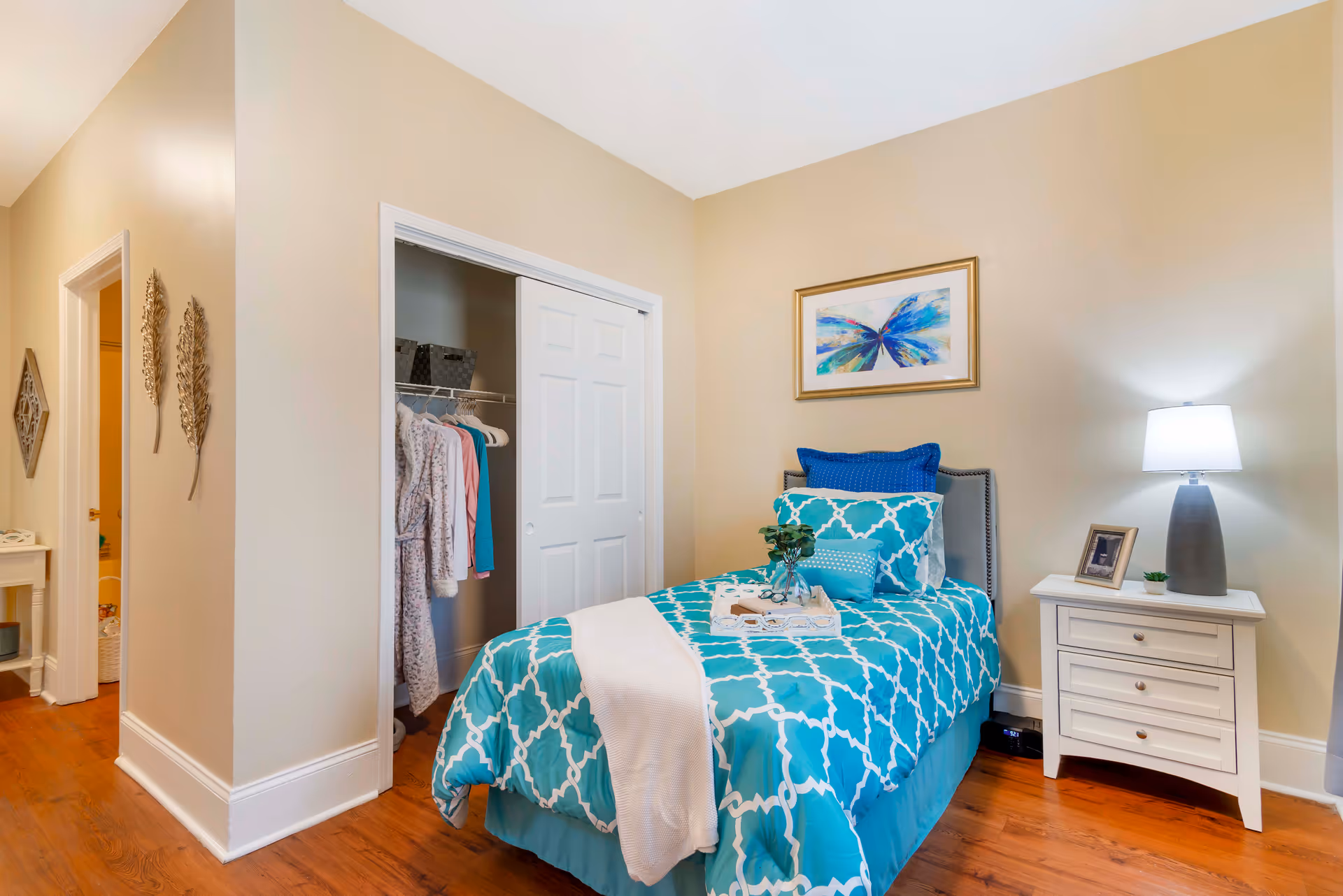 A cozy bedroom with a single bed dressed in turquoise and white patterned bedding, a white nightstand with a lamp and picture frame, an open closet with clothes hanging inside, and beige walls adorned with a colorful butterfly painting and decorative wall hangings.