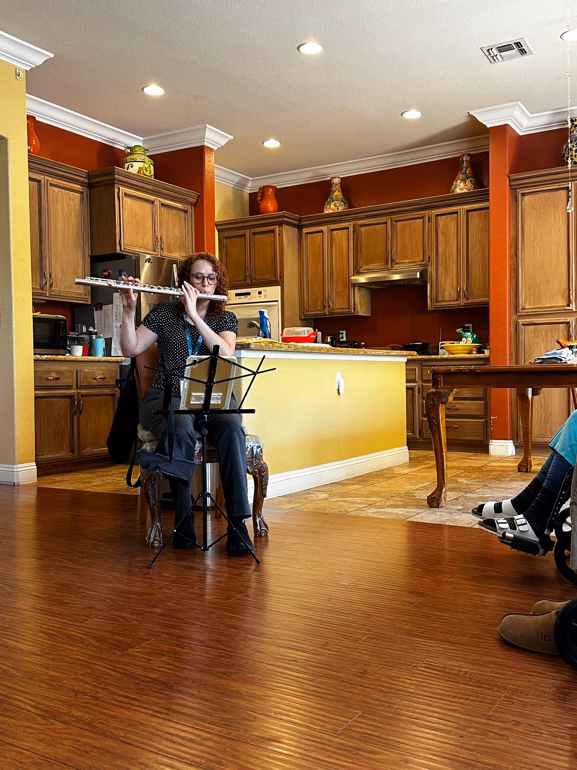 A woman with curly red hair and glasses is sitting on a chair playing the flute in a kitchen area with wooden cabinets and a yellow island. There is a music stand in front of her, and part of a person in a wheelchair is visible on the right side of the image.