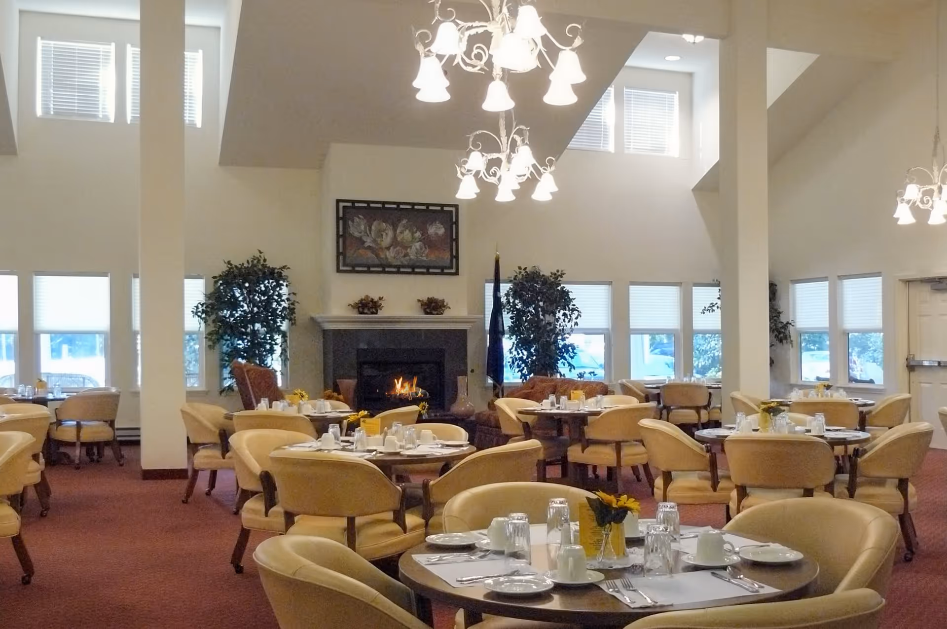 A spacious dining room with round tables set for meals, beige cushioned chairs, a lit fireplace in the background, large windows with blinds, and chandeliers hanging from a high ceiling.