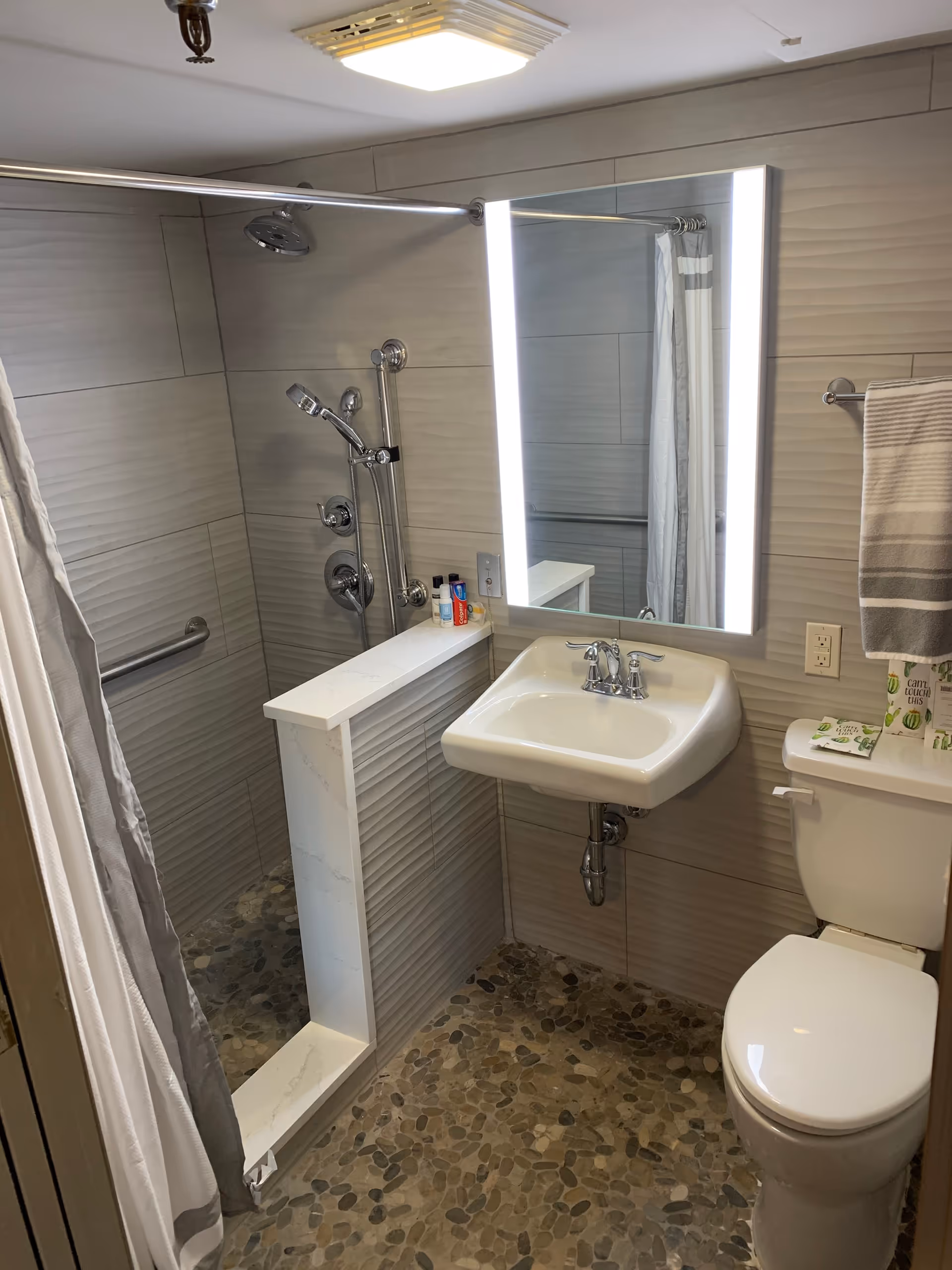 A modern bathroom with a walk-in shower featuring a handheld and overhead showerhead, a white sink with chrome faucet, a large illuminated mirror, a toilet with a towel rack above it holding a striped towel, and a pebble stone floor.