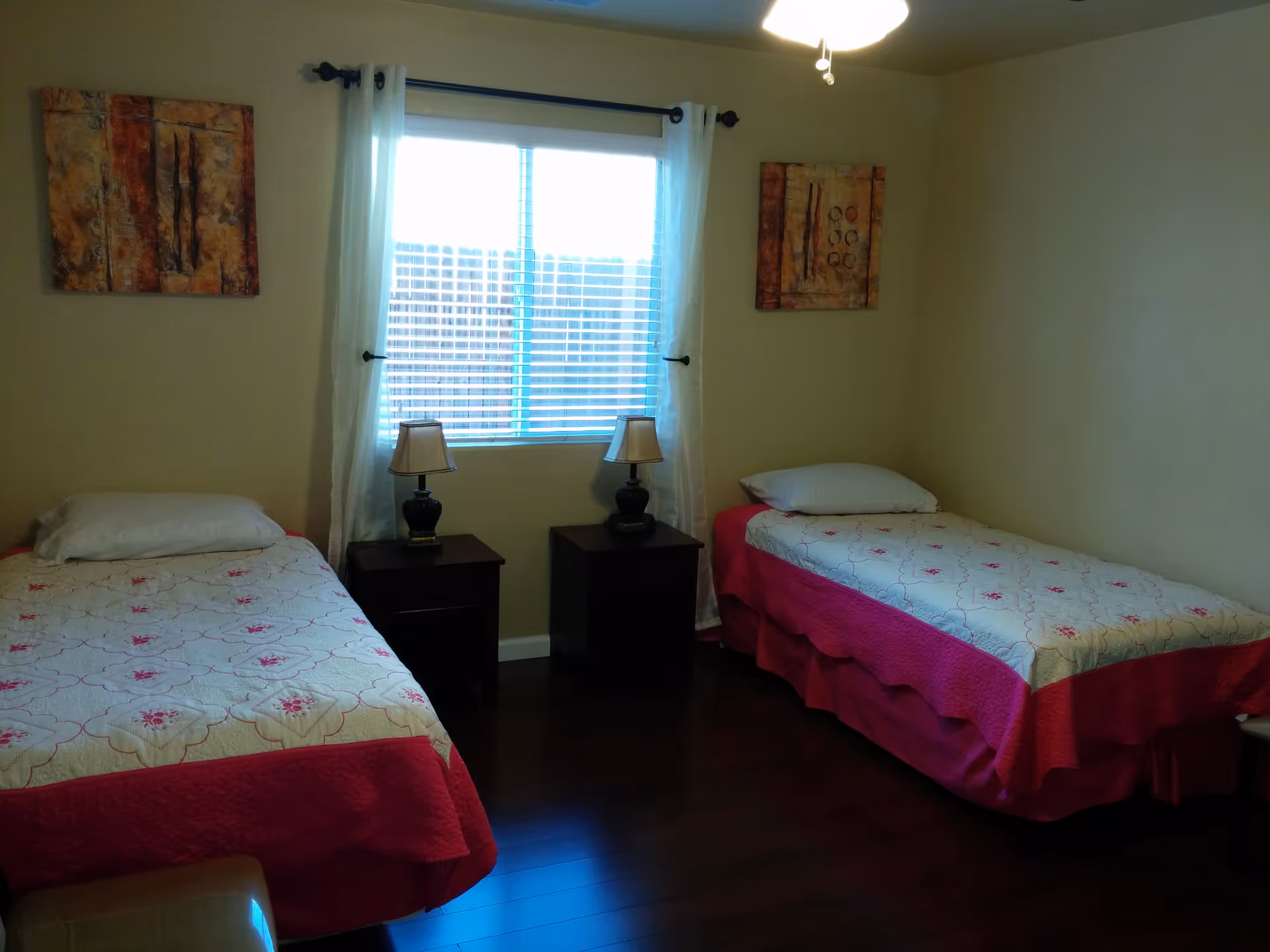 A bedroom with two single beds placed against opposite walls, each bed covered with a white and pink patterned bedspread and a pillow. Between the beds are two dark wooden nightstands, each with a lamp. A window with white curtains is centered on the wall behind the nightstands. Two abstract paintings hang on the walls above each bed. The room has wooden flooring and a ceiling fan with lights.