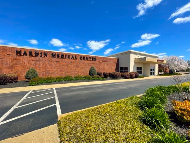 Exterior view of Hardin Medical Center building with a brick facade and clear blue sky. There is a driveway and landscaped greenery with bushes and grass in front of the building.