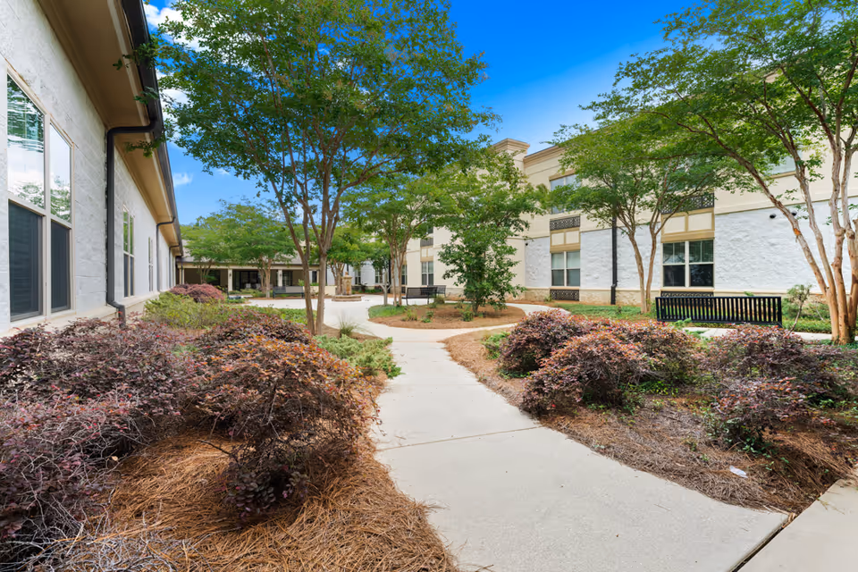 Outdoor courtyard area at Seabrook Pointe featuring a paved walkway surrounded by landscaped bushes and trees, with benches along the path and the building exterior visible under a clear blue sky.