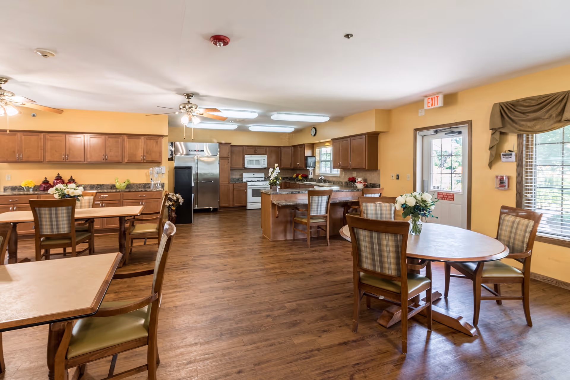 A spacious senior living facility dining area with wooden floors, multiple tables and chairs, and a large kitchen in the background featuring wooden cabinets, stainless steel refrigerator, microwave, and stove. The room is well-lit with ceiling fans and fluorescent lights, and there are windows with blinds and a door leading outside.