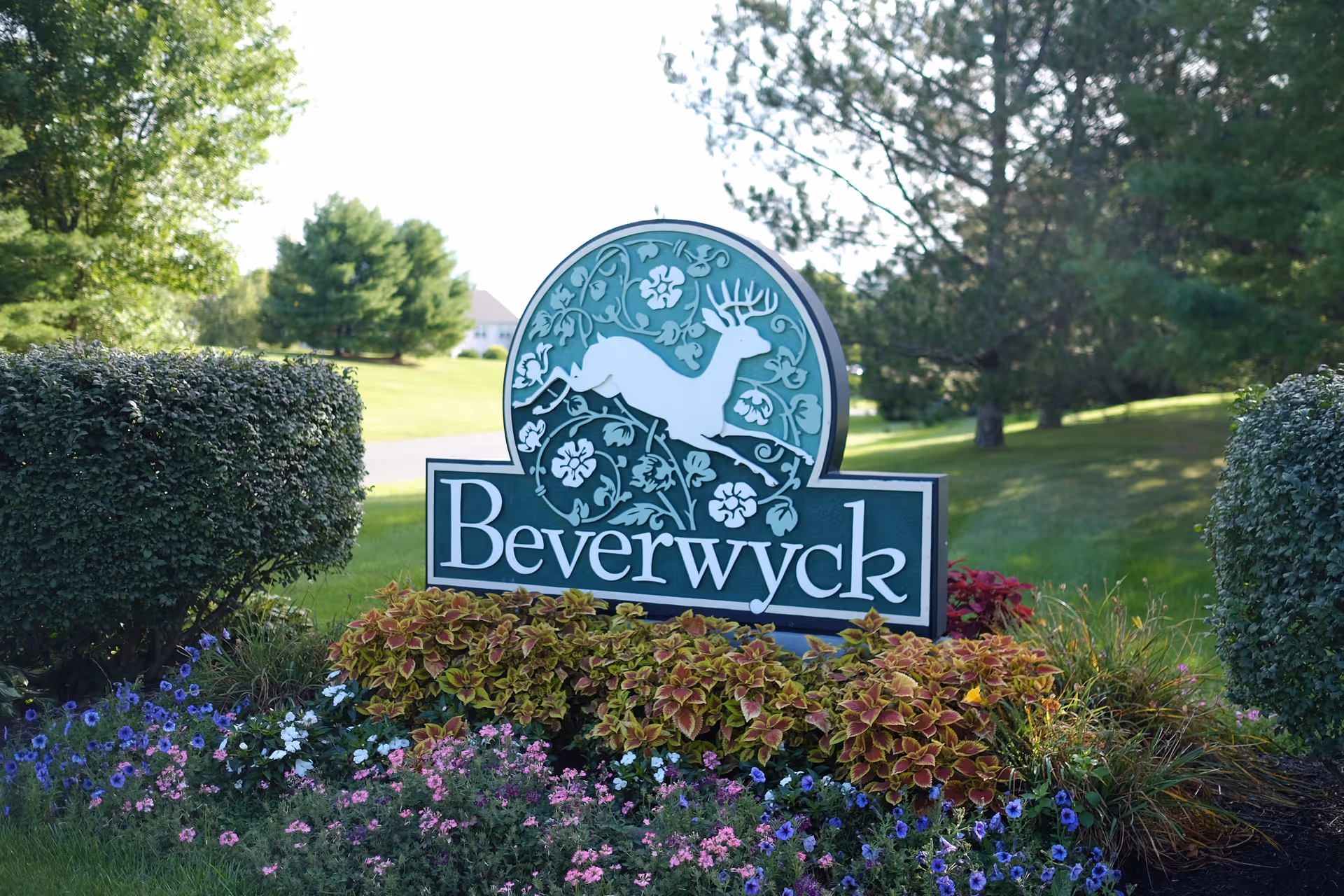 A green and white sign with a leaping deer and floral design that reads 'Beverwyck' surrounded by colorful flowers and bushes in a grassy outdoor area with trees in the background.