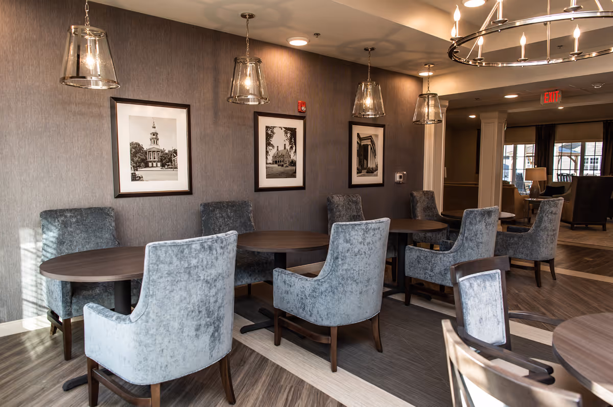 A modern communal dining area with round wooden tables, upholstered gray chairs, pendant lights, and framed black-and-white photos on the wall.