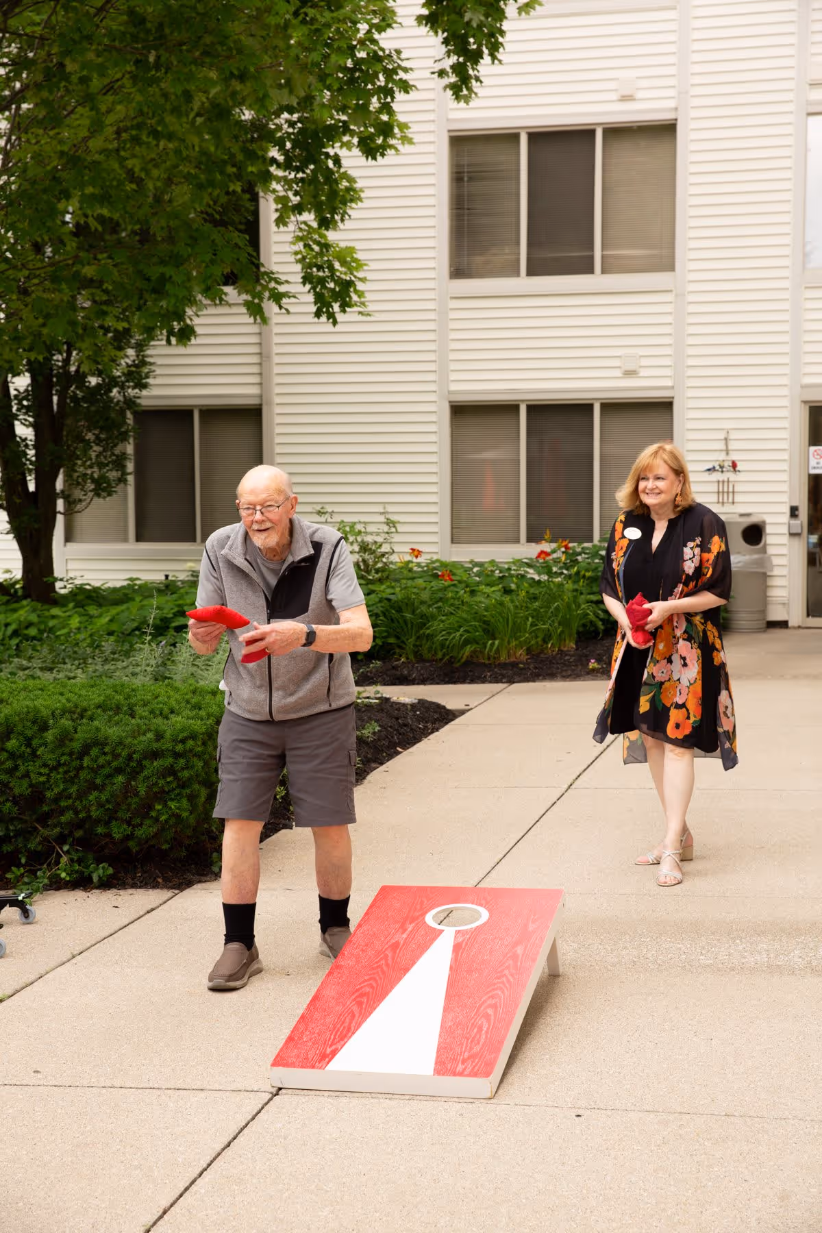 An elderly man and a woman playing cornhole outside on a paved area in front of a white building with windows and greenery. The man is in mid-throw holding a red bean bag, while the woman stands behind him holding another red bean bag, smiling.
