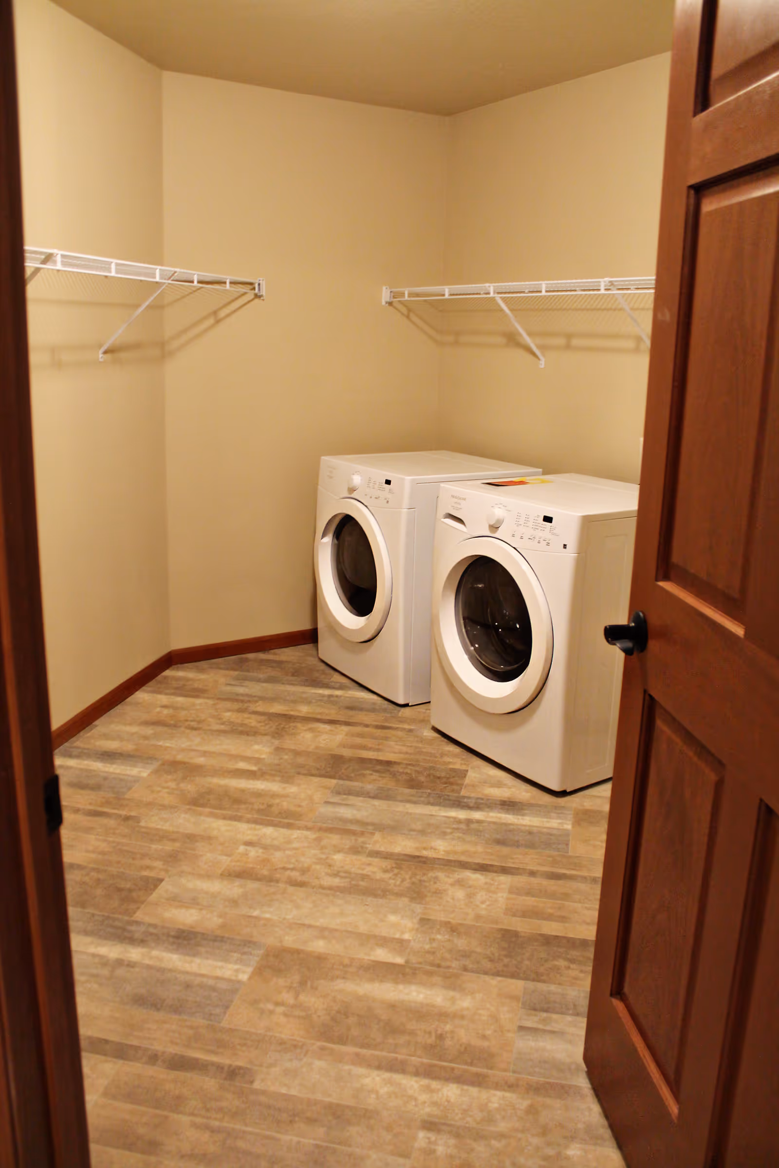 Laundry room with a front-loading washing machine and dryer side by side, beige walls, wood trim, and wire shelving on two walls.