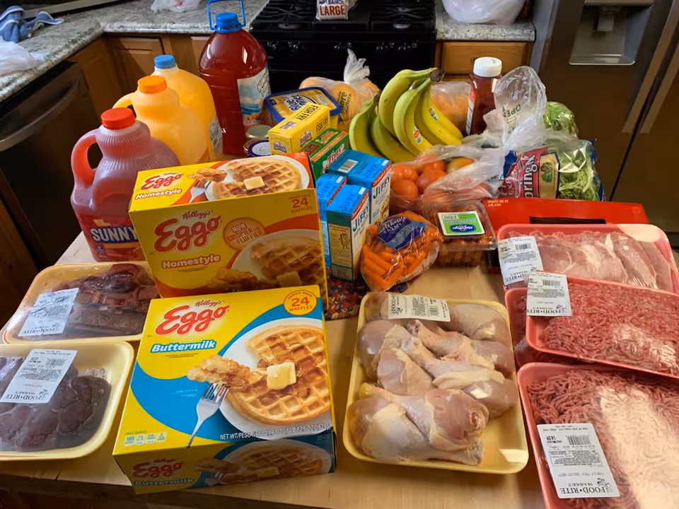 A kitchen countertop filled with various groceries including packs of raw meat, boxes of Eggo waffles, cartons of Jiffy corn muffin mix, bags of fresh produce such as bananas, tomatoes, carrots, lettuce, and bottles of juice and ketchup.