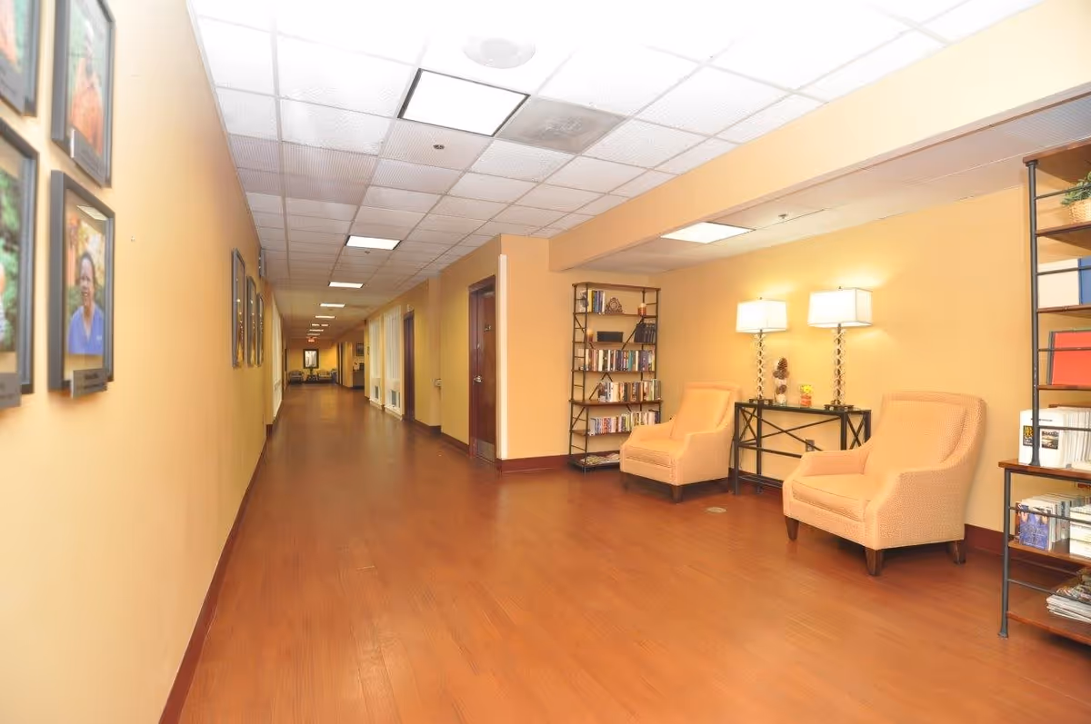 Long, well-lit interior hallway with seating area, bookshelves, framed photos, and wood flooring.