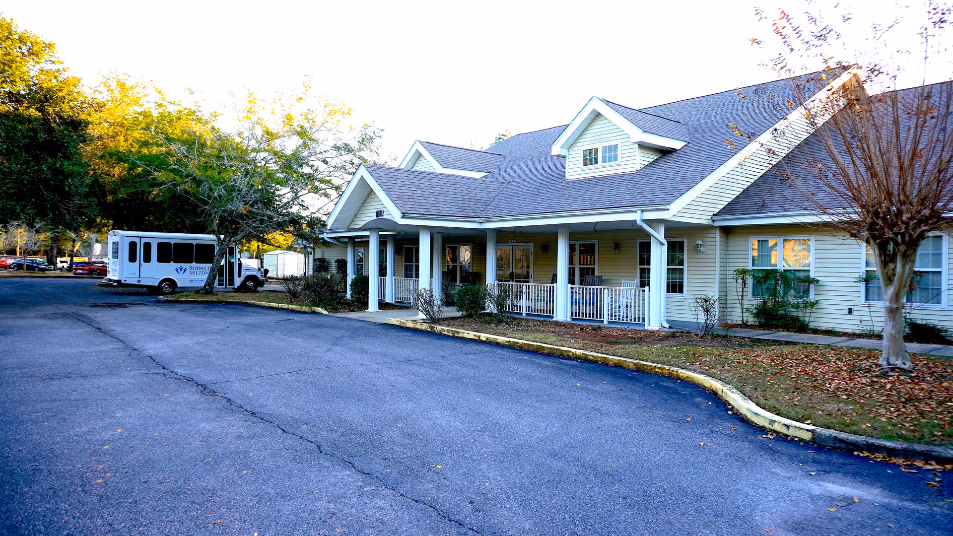 Front exterior of a one-story senior living building with a covered porch, parking lot, and a shuttle bus parked to the left.