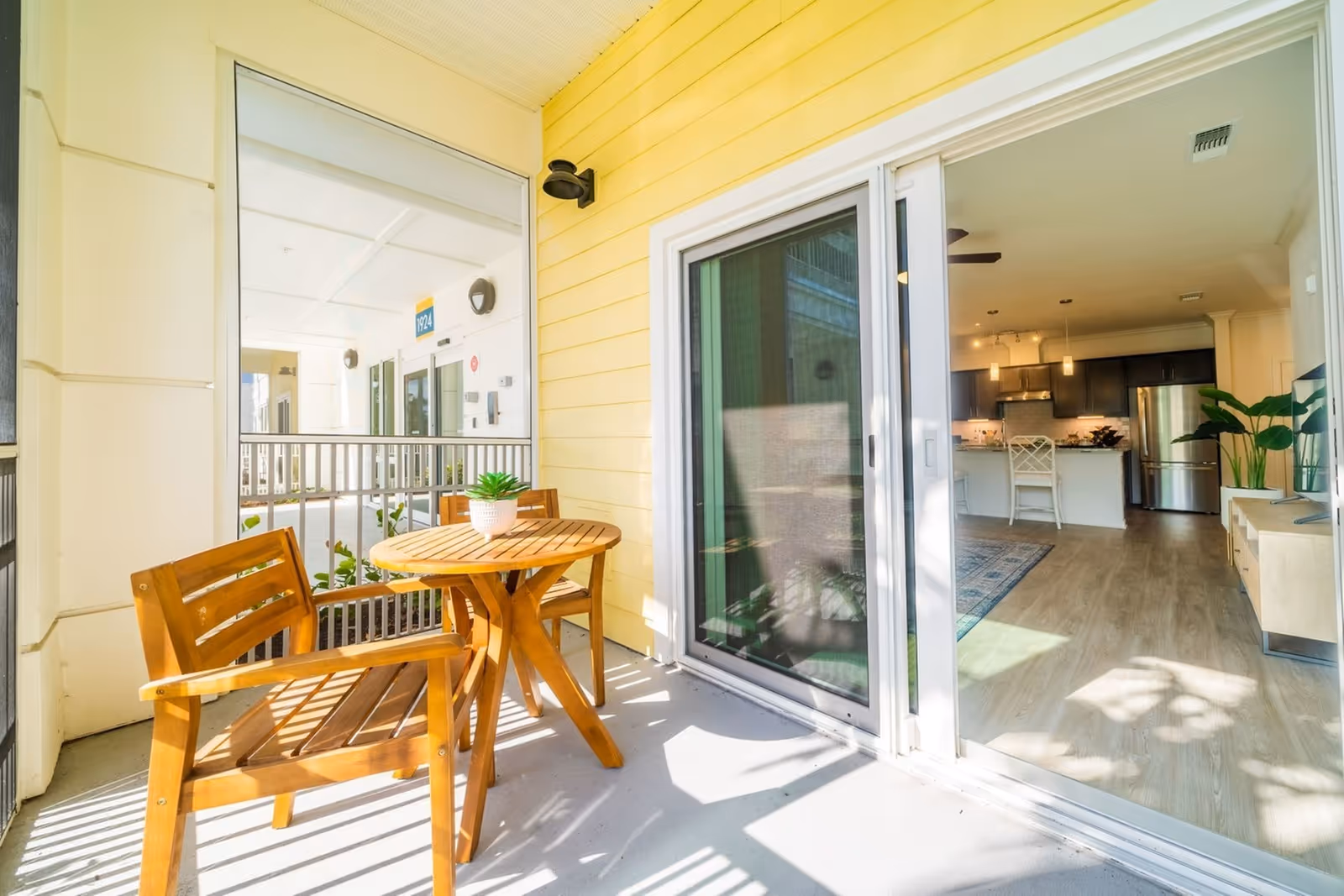 A sunlit balcony with a wooden table, chairs and potted plant opening through sliding glass doors to a modern kitchen and living area.