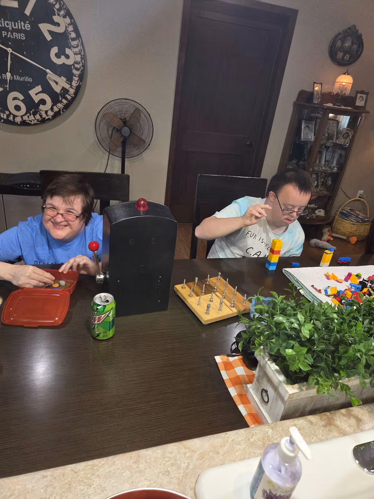 Two individuals sitting at a dining table engaged in activities. One person is smiling and appears to be playing a game with tokens, while the other is stacking colorful building blocks. The table has a can of Mountain Dew, a game board with pegs, and a decorative plant. In the background, there is a large wall clock, a standing fan, a wooden door, and a cabinet with framed photos and decorative items.
