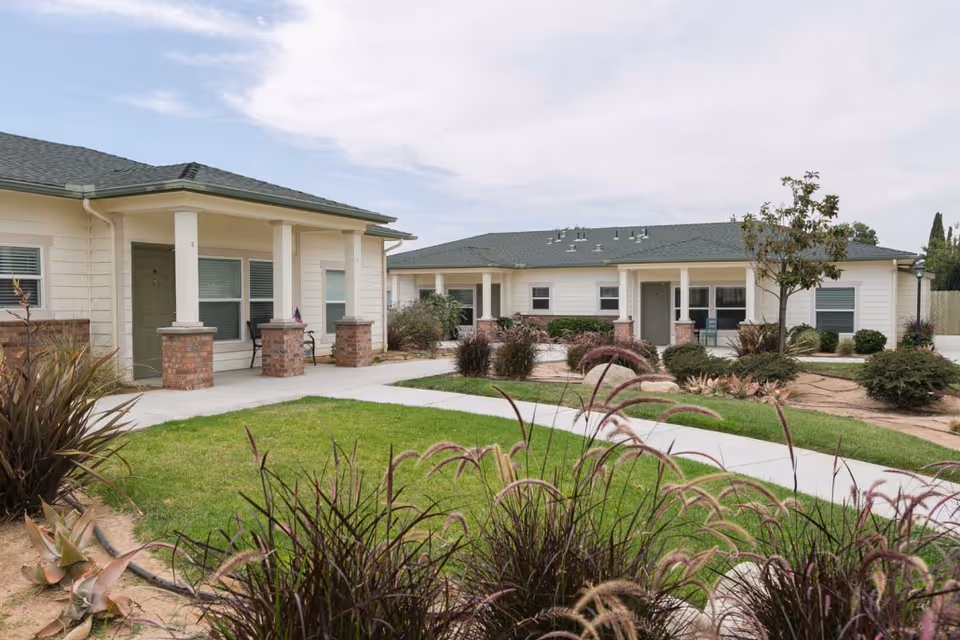 Outdoor view of a senior living facility with single-story buildings featuring covered porches, brick pillars, and green lawns with landscaped plants and walking paths under a partly cloudy sky.