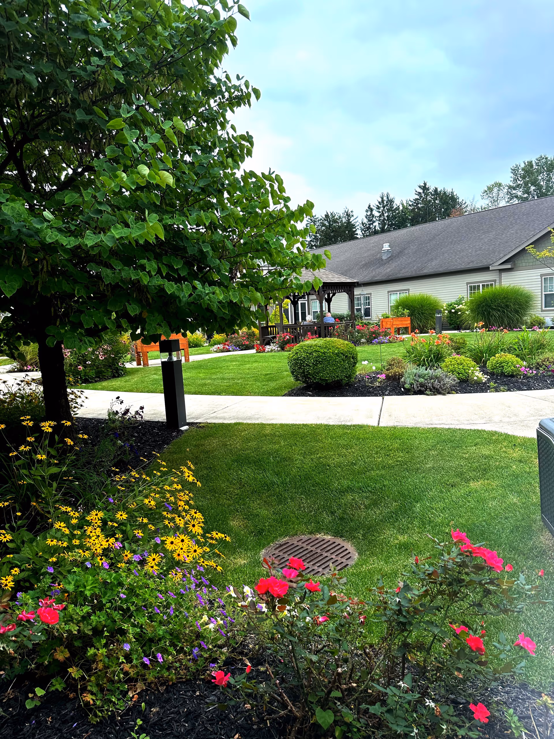 A well-maintained outdoor garden area with green grass, colorful flowers including yellow and red blooms, a large leafy tree, and a paved walkway. In the background, there is a gazebo with people sitting inside and a single-story building with a gray roof and light-colored siding.