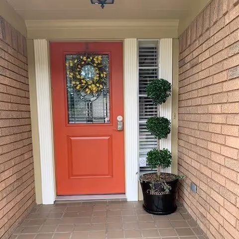 Red front door with wreath and potted topiary beside brick entryway.