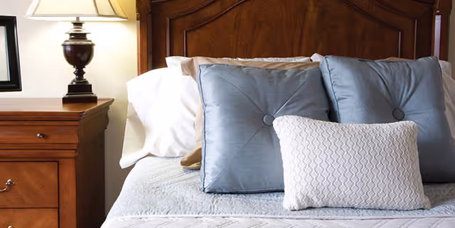 Close-up view of a neatly made bed with two blue decorative pillows and one white textured pillow, next to a wooden nightstand with a lamp and a small part of a television visible.