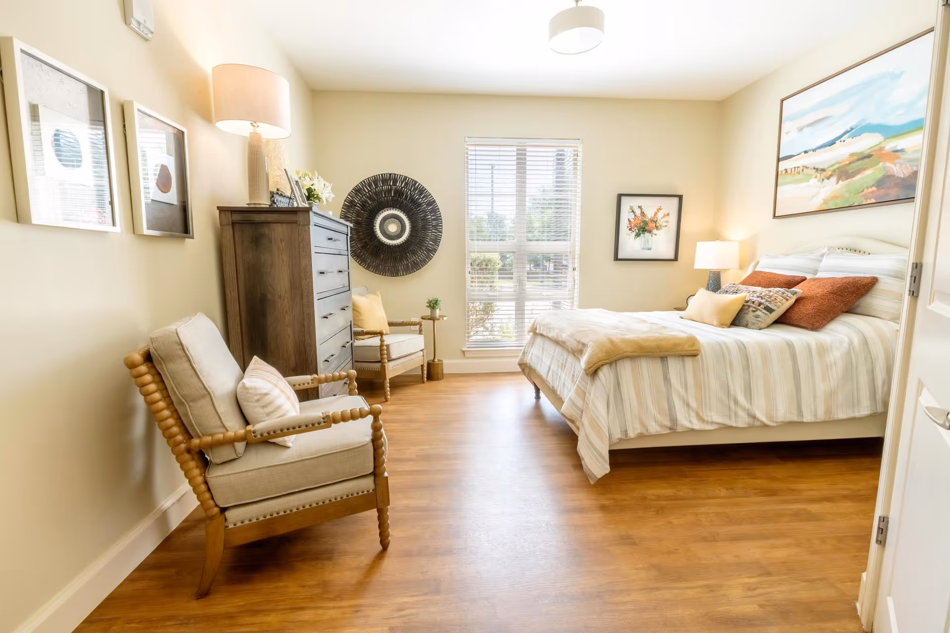 Bright, tastefully furnished bedroom with a bed, two armchairs, a dresser, and a window letting in natural light.