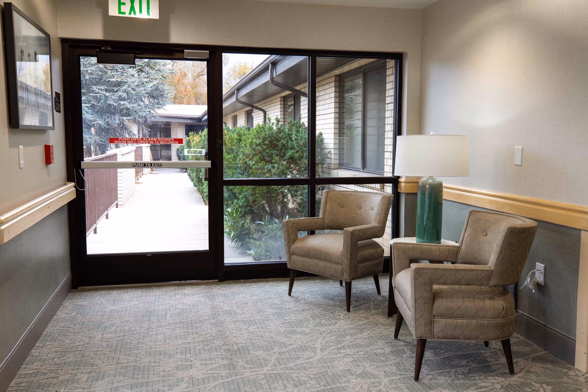 A small seating area inside a facility with two beige upholstered armchairs and a side table with a green lamp. The area is next to a glass door and window that show an outdoor walkway with bushes and trees. An exit sign is visible above the door.