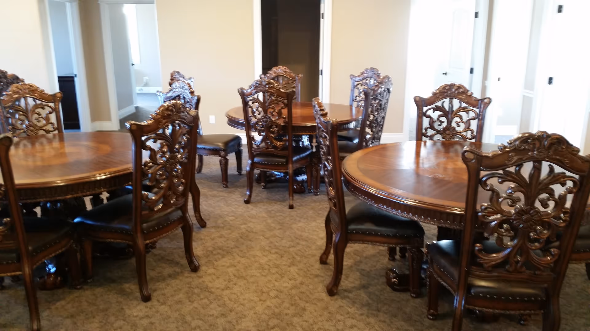 Interior view of a dining room with multiple round wooden tables and intricately carved wooden chairs with dark cushioned seats. The room has beige walls and carpeted floor, with doorways visible in the background.