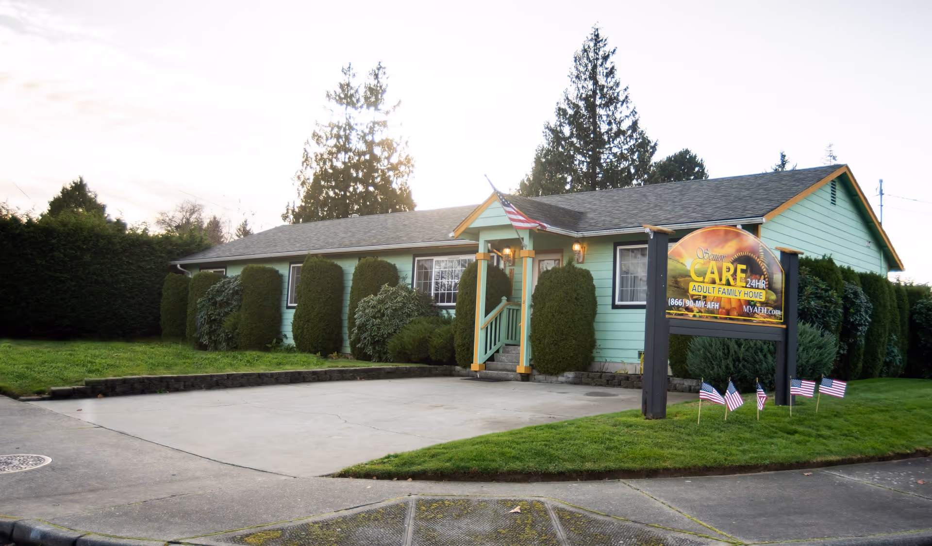 Single-story mint-green senior care home with a front porch, landscaped shrubs, a driveway, American flags, and a large sign by the lawn.