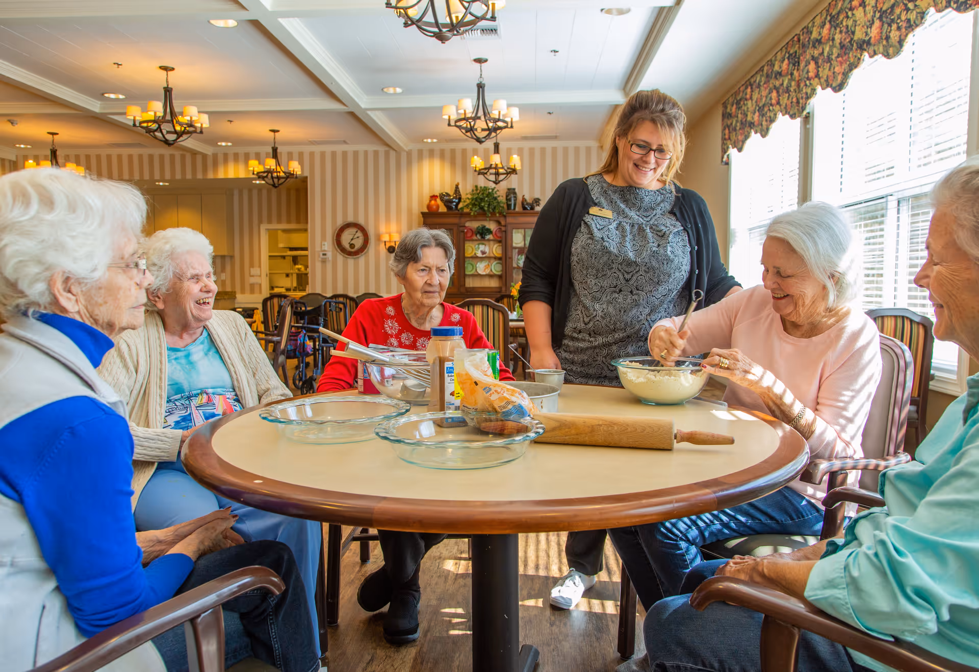A group of elderly women sitting around a round table in a well-lit room with large windows and floral curtains. They are engaged in a baking activity with mixing bowls, a rolling pin, and ingredients on the table. A caregiver or staff member stands smiling beside them, observing and assisting. The room has warm lighting with chandeliers and striped wallpaper.
