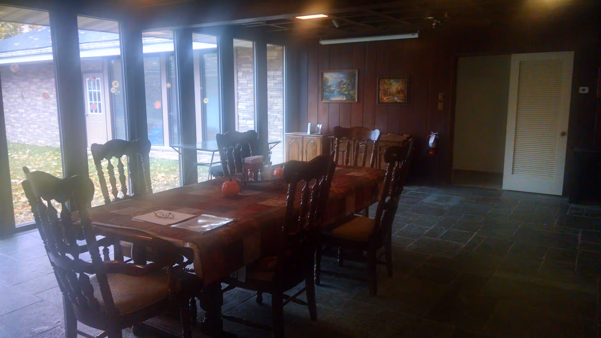 Interior view of a dining room with a long wooden table covered with a patterned tablecloth and surrounded by eight wooden chairs. Large windows on one side let in natural light and show an outdoor area with grass and a building. The room has dark wood-paneled walls with two framed paintings and a small cabinet. The floor is tiled with large dark tiles, and there is an open door leading to another room.