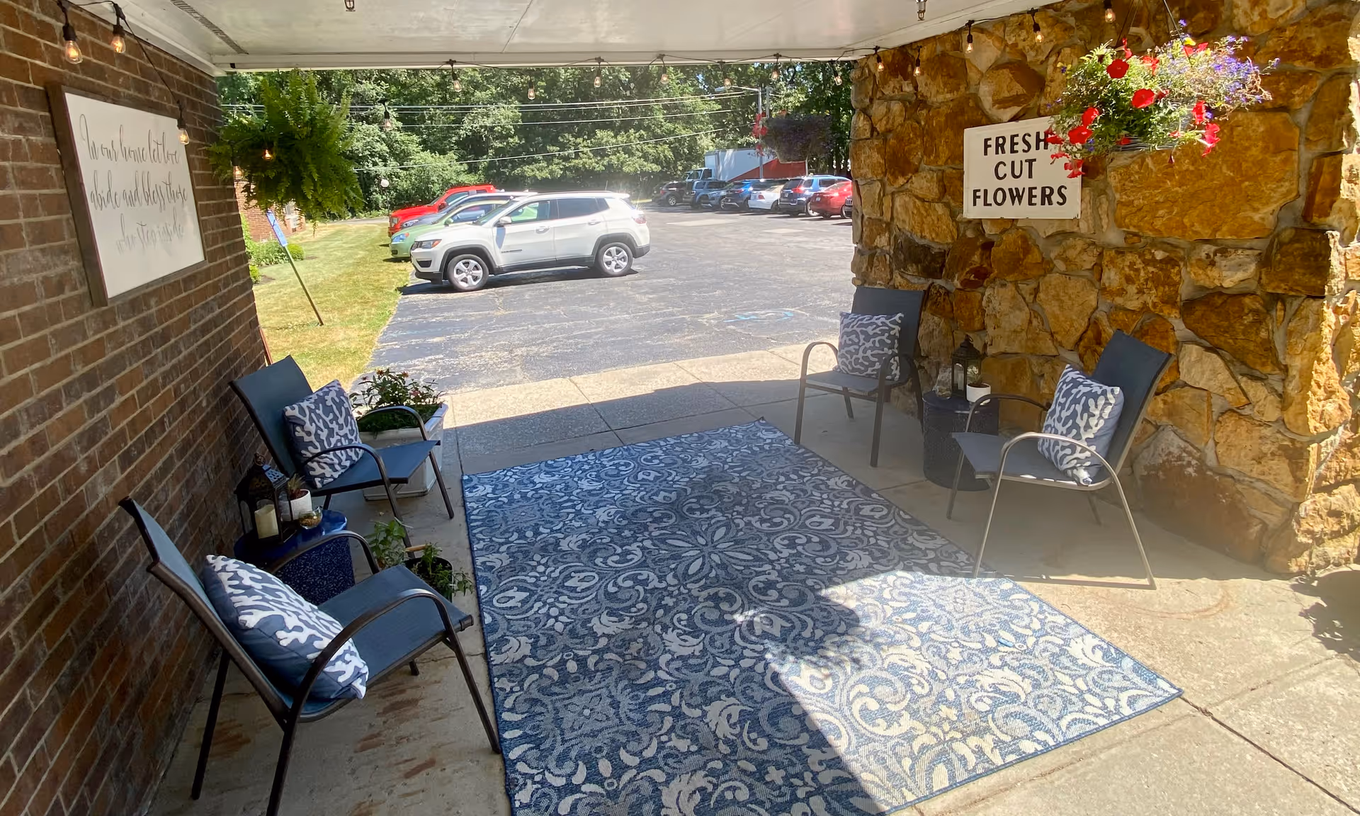 Covered outdoor seating area with four chairs, each with patterned cushions, arranged around a blue and white patterned rug. There are hanging plants and a sign on a stone wall that reads 'FRESH CUT FLOWERS'. In the background, a parking lot with several cars is visible.