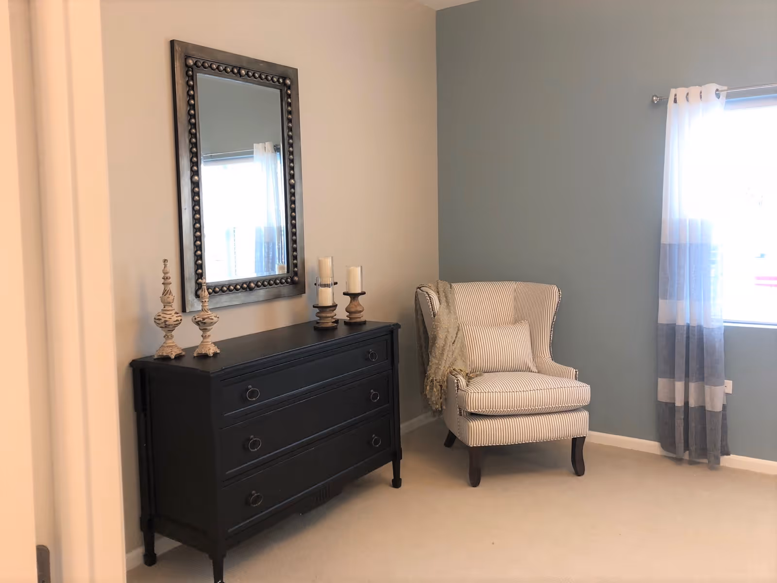 A cozy room corner with a black dresser topped by decorative candlesticks and a mirror, beside a striped wingback chair next to a curtained window.