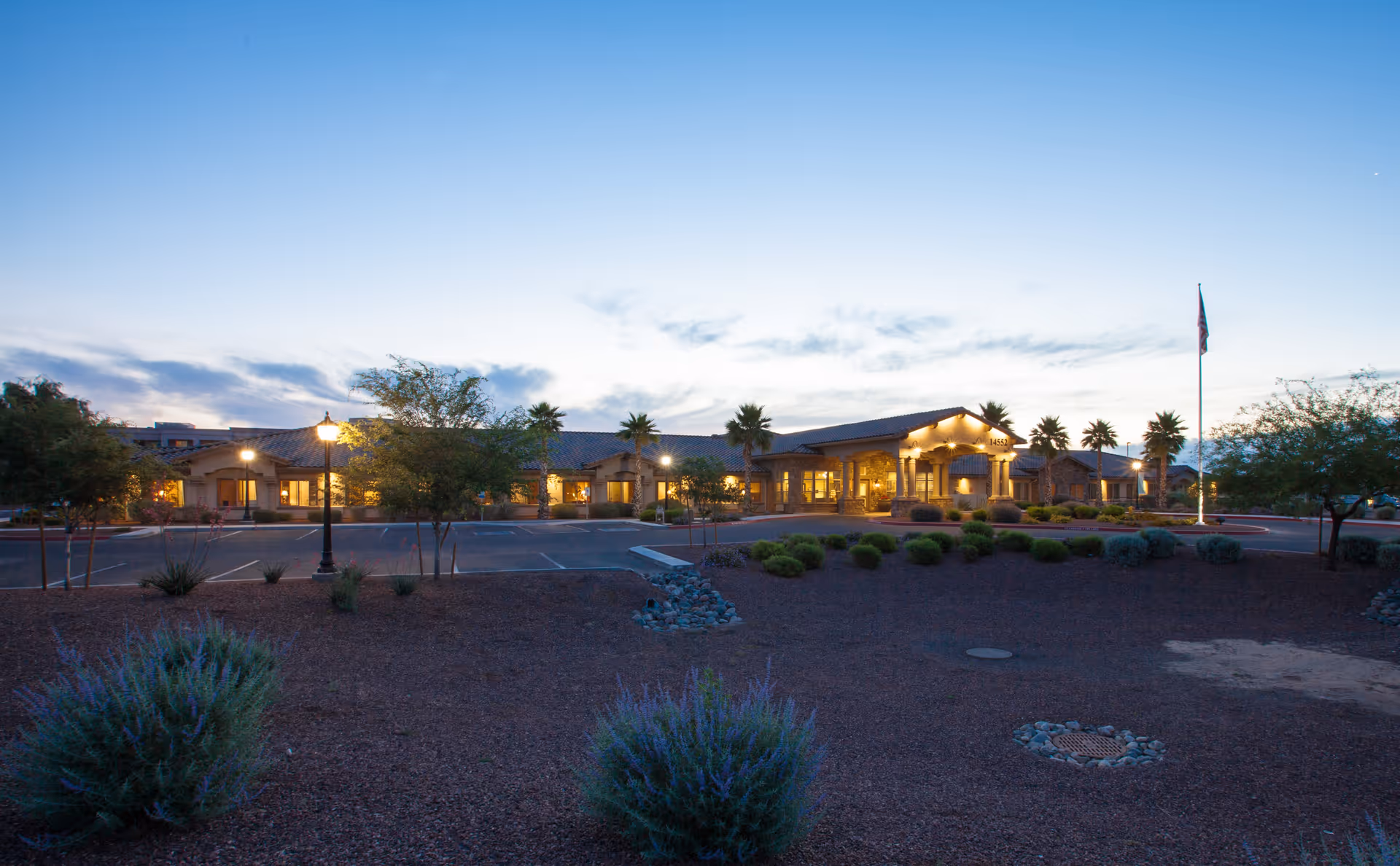 Dusk exterior view of a single-story memory care building lit from inside, with a parking lot, desert landscaping, palm trees, and a flagpole.