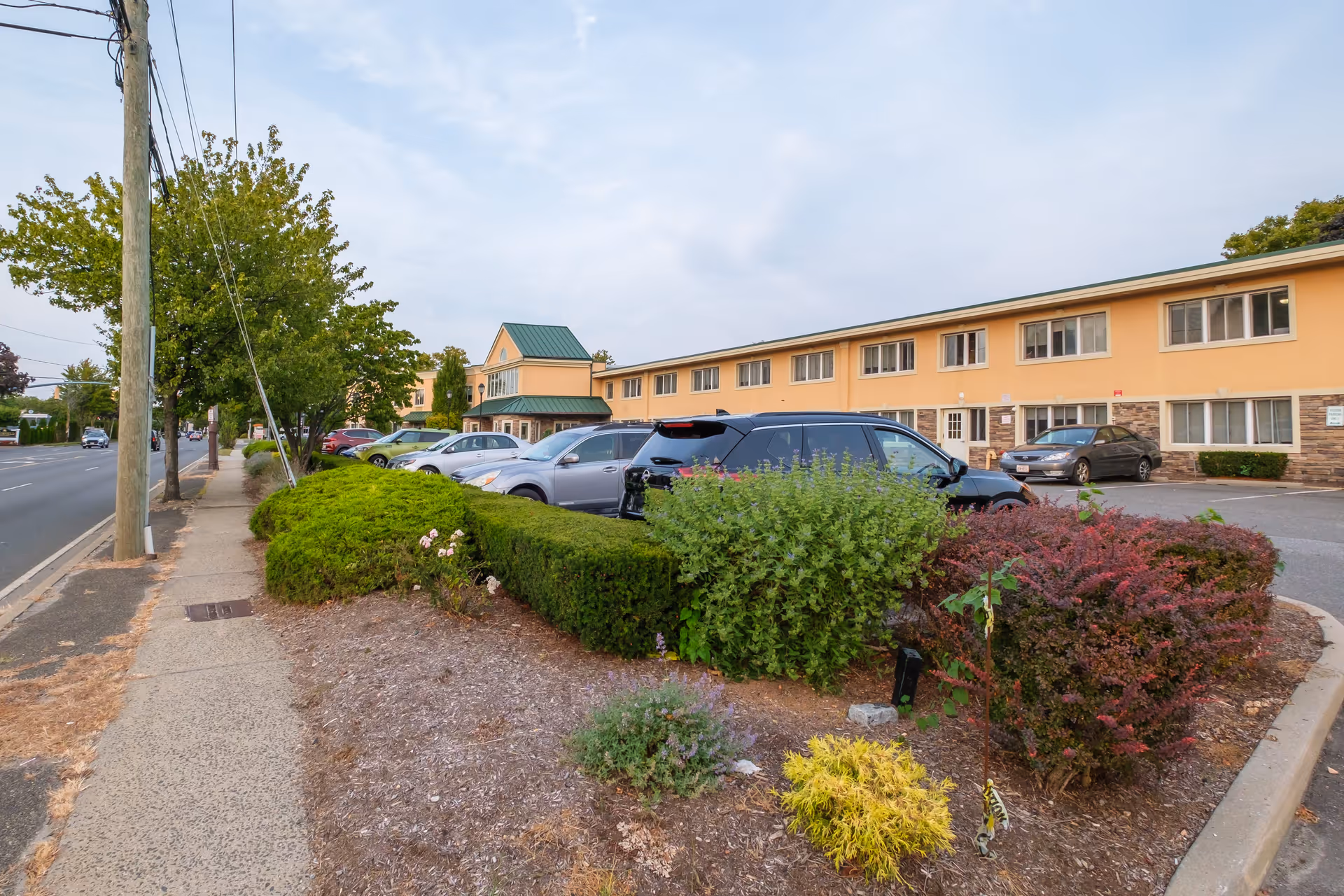 Exterior view of Amber Court of Westbury, showing a two-story building with beige walls and green roof accents. Several cars are parked in the parking lot in front of the building, and there are bushes and small plants along the sidewalk and parking area. A sidewalk runs alongside the street with trees and utility poles visible.