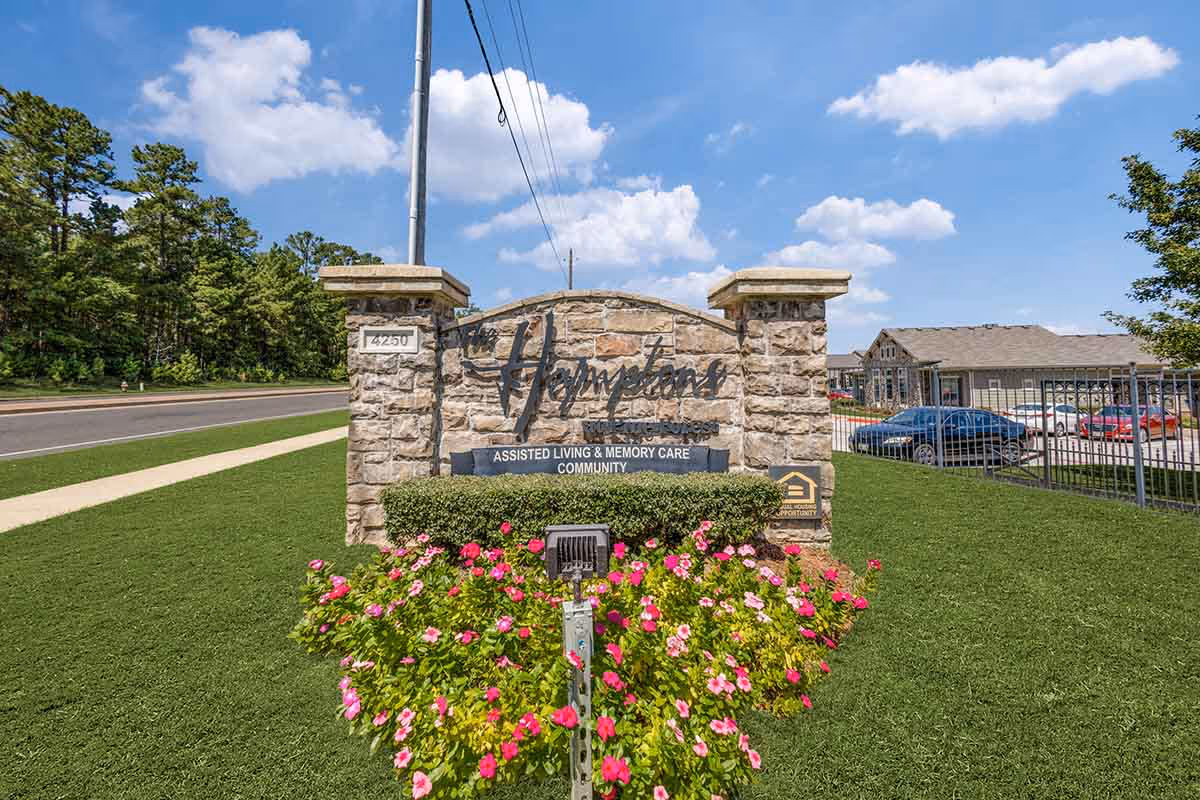 Stone sign for The Hamptons Of Tyler assisted living and memory care community, surrounded by green grass and pink flowers, with a road and trees in the background under a blue sky with clouds.