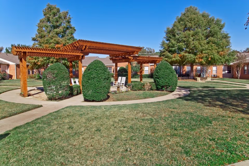Sunlit courtyard featuring a wooden pergola, trimmed shrubs, walkways, and surrounding red-brick residential buildings.