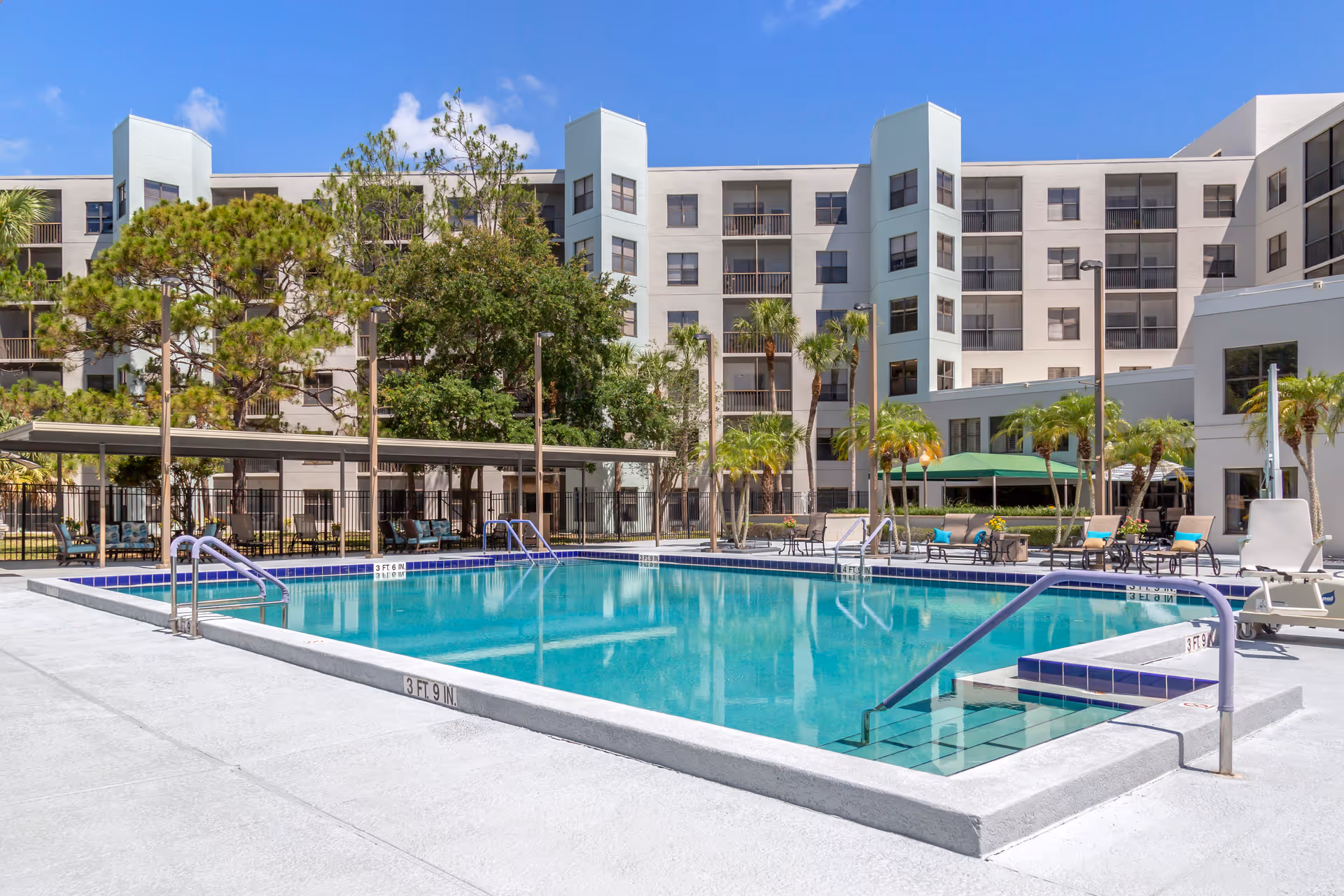 Outdoor swimming pool with clear blue water surrounded by a concrete deck, lounge chairs, palm trees, and a multi-story residential building in the background under a clear blue sky.