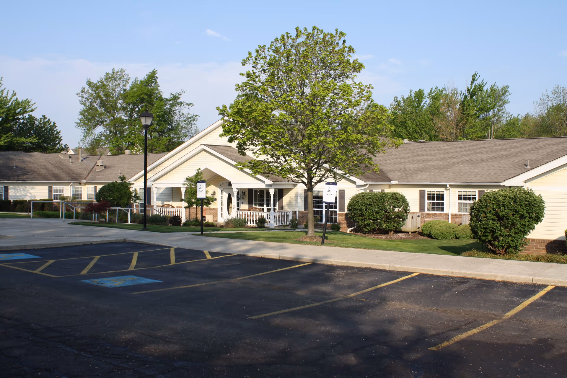 Exterior view of Arden Courts - ProMedica Memory Care Community (Westlake) building with a parking lot in front, including handicapped parking spaces, a tree, bushes, and a lamppost under a clear blue sky.