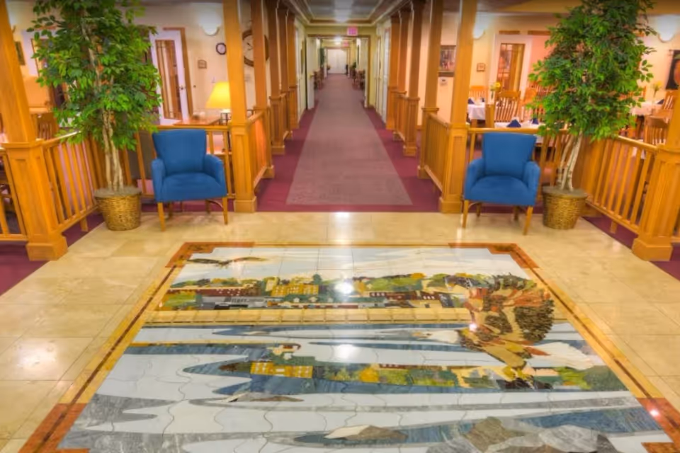 Interior hallway of a senior living facility with a decorative floor mural depicting a nature scene, flanked by two blue armchairs and potted plants. The hallway has wooden railings and columns, with a carpeted walkway leading to rooms and a dining area visible on the right.