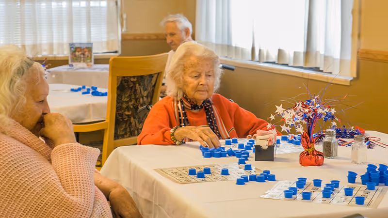 Three elderly individuals sitting at tables in a well-lit room playing a game of bingo with blue markers on bingo cards. The tables are covered with white tablecloths and decorated with a red, white, and blue centerpiece. The room has beige walls and windows with white curtains.