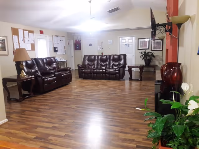 A spacious common area with wooden flooring featuring two dark brown leather sofas, a side table with a lamp, a wall-mounted TV, potted plants, framed pictures on the walls, and a bulletin board near the entrance doors.
