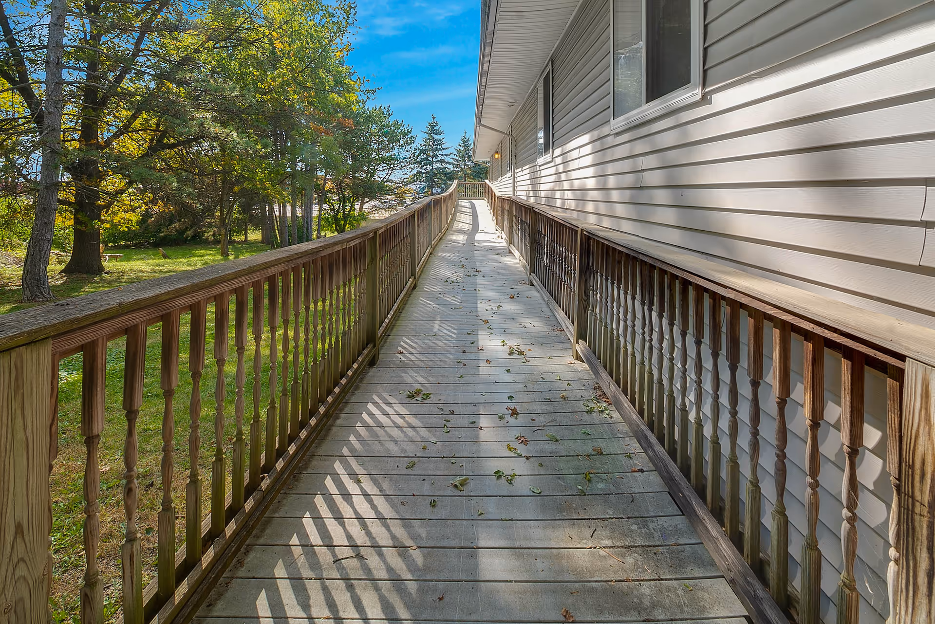 A wooden outdoor ramp with railings on both sides leading up alongside a building with white siding. Trees and green grass are visible on the left side under a clear blue sky.