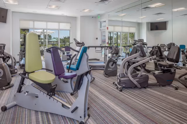 Bright fitness room with recumbent cardio machines, strength equipment, a mirrored wall and windows overlooking an outdoor area.