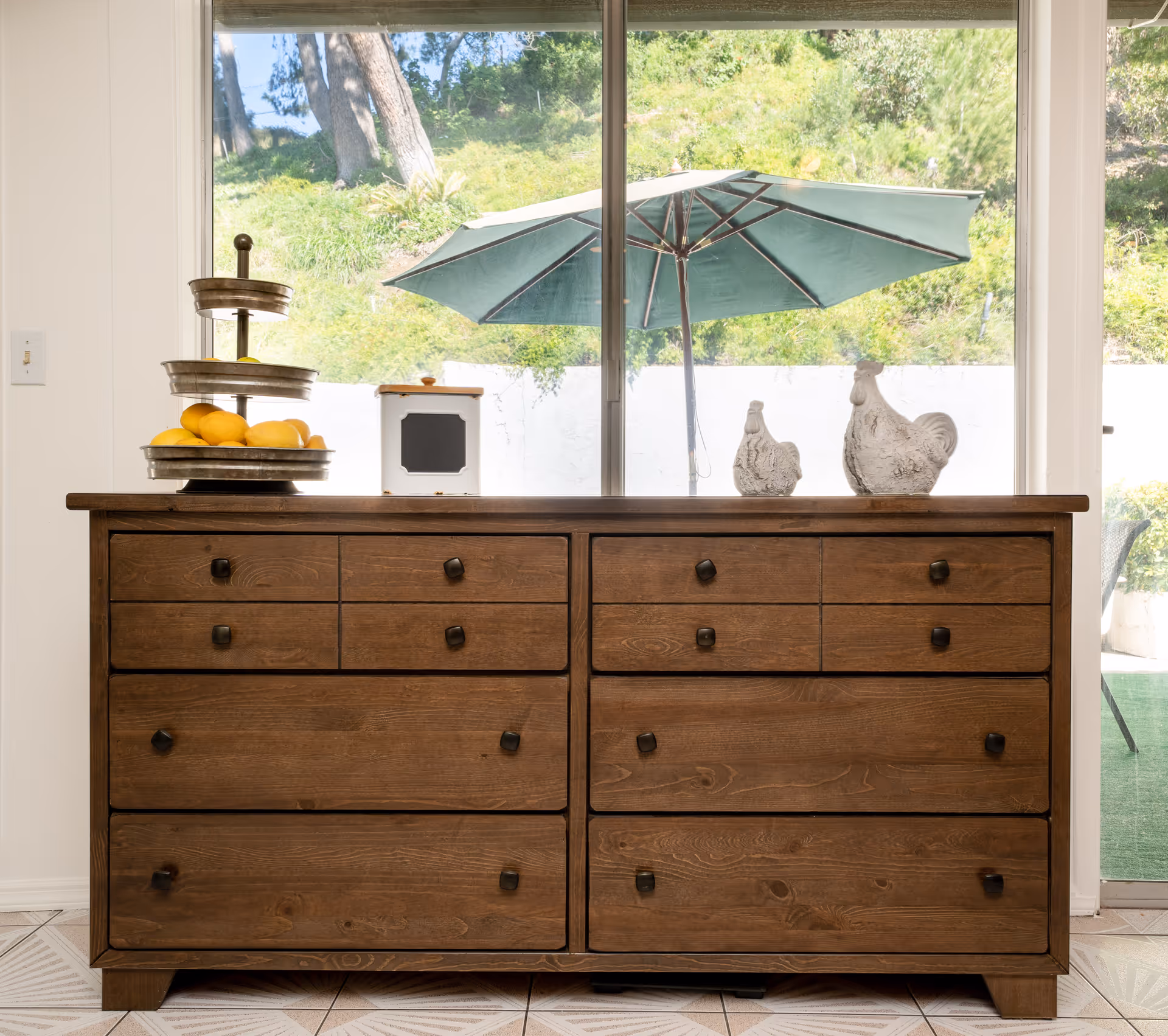 Wooden dresser topped with decorative items in front of a sliding glass door showing a patio umbrella and greenery outside.