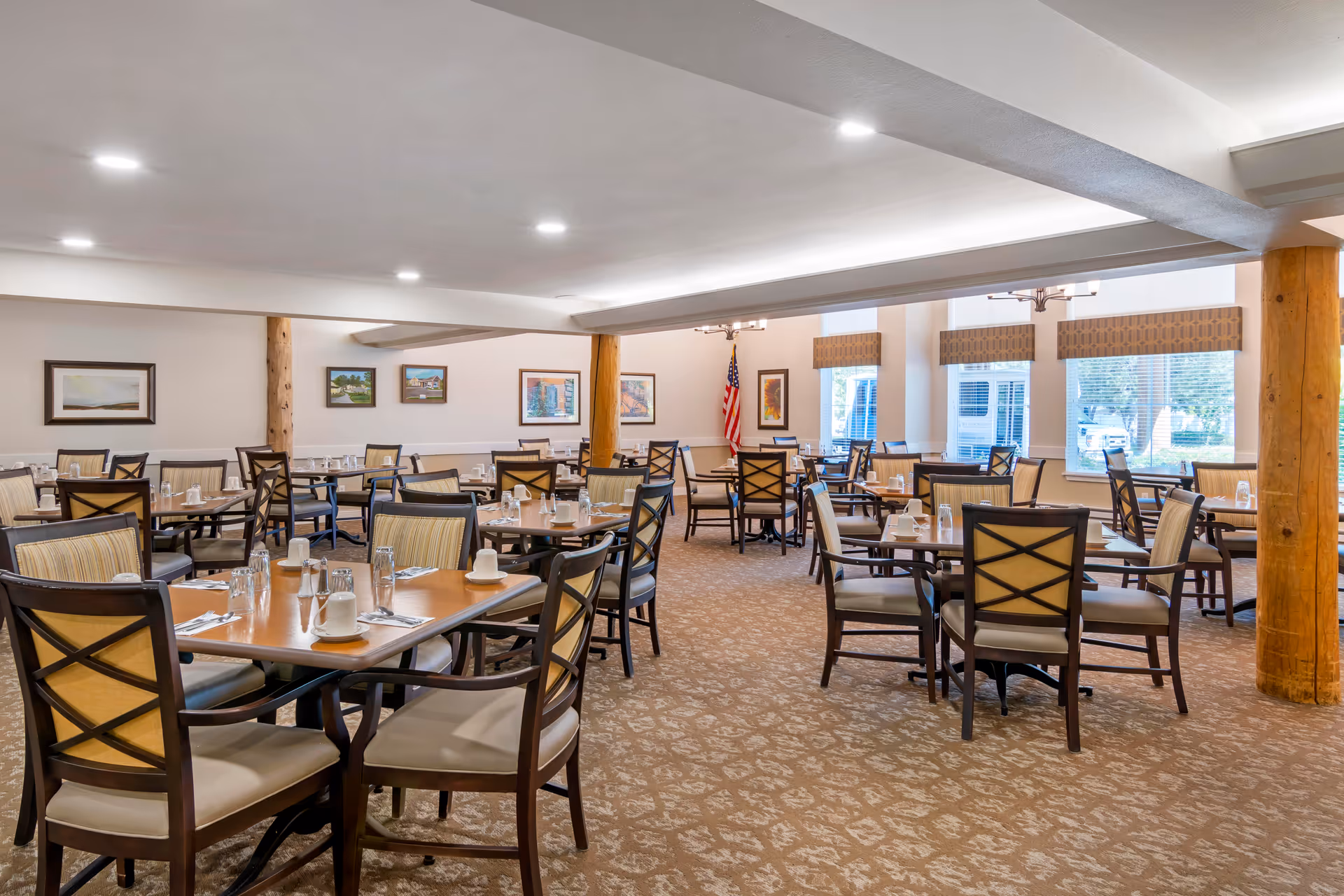 A spacious dining room with multiple wooden tables and cushioned chairs arranged neatly. Each table is set with cups, glasses, silverware, and napkins. The room has large windows with valances, framed artwork on the walls, and an American flag in the corner. The carpet has a patterned design, and wooden support beams are visible.