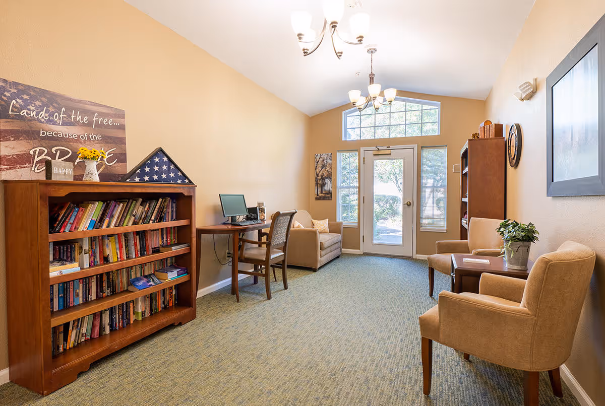 A cozy senior living common area with a bookshelf filled with books, a small desk with a computer, two armchairs with a small table between them, and a loveseat near a glass door with windows letting in natural light. The walls are beige, and there is a patriotic sign on the bookshelf that reads 'Land of the free... because of the brave.'
