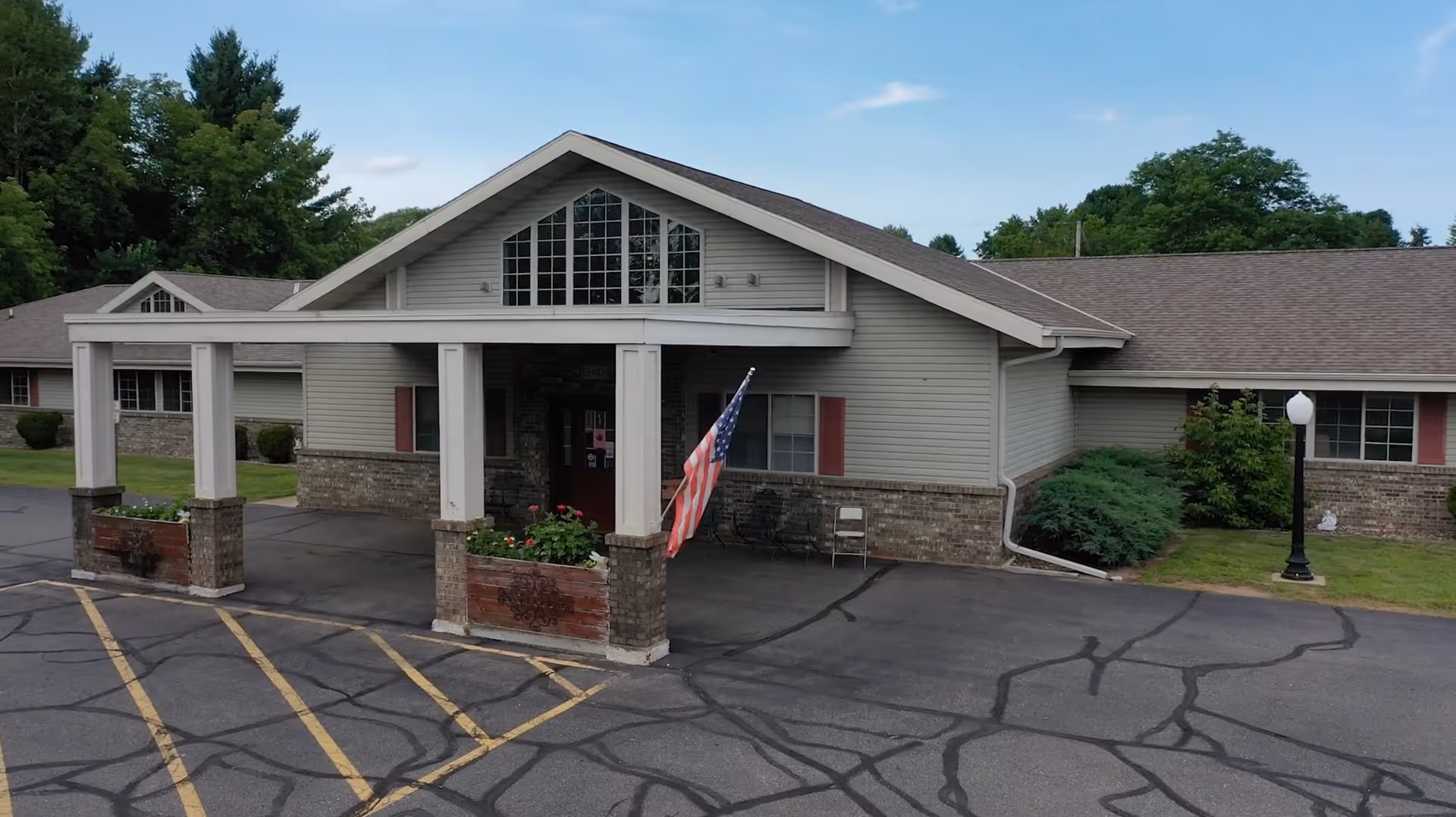 Front exterior view of a single-story senior living facility building with a covered entrance supported by white columns and brick planters. An American flag is displayed near the entrance, and there are parking spaces with yellow lines in front. The building has beige siding, brick accents, and multiple windows with red shutters. Trees and greenery surround the area under a blue sky with some clouds.